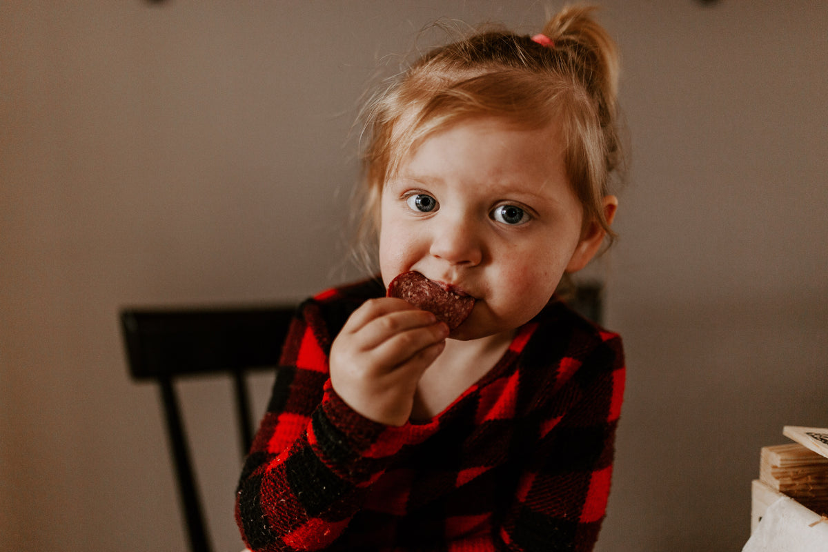 A little girl in a plaid dress enjoying a slice of summer sausage